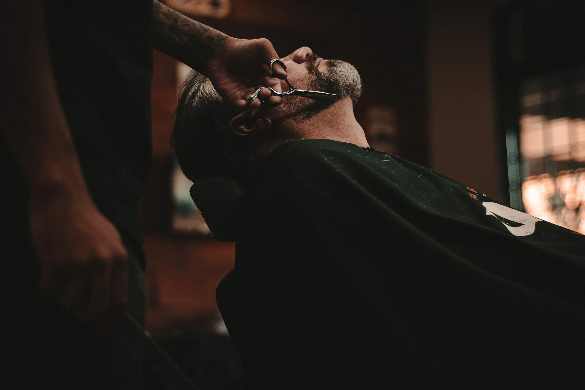 Ein Barbier mit tätowiertem Unterarm stutzt einem zurückgelehnten Kunden mit der Schere den grau melierten Bart.